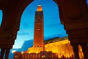 A large, illuminated mosque with a tall minaret at twilight, framed by an ornate archway