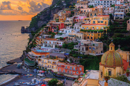 Colorful buildings perched on a steep hillside overlook the ocean during sunset, with boats docked along the shoreline below